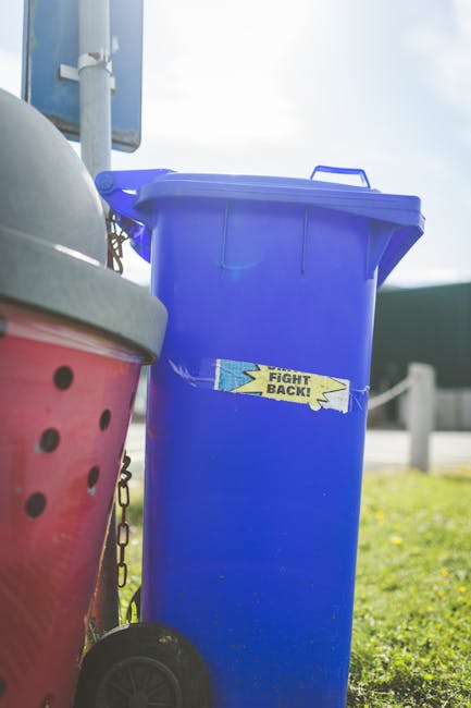 A tall, closed blue wheelie bin made of smooth plastic with a slightly textured surface, positioned outdoors on a grassy area near a paved surface. The bin has a matching blue lid with a handle at the