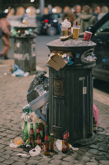 A mound of mixed waste materials and household rubbish on a gravel surface outdoors, including black plastic bags, a yellow plastic container, and the remains of an old, dirty car seat or upholstery f